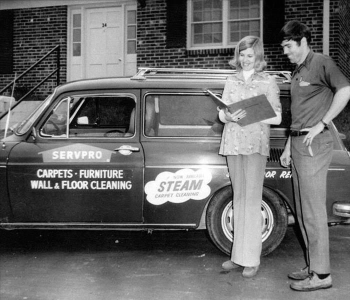 A historic black and white photo of a SERVPRO professional and customer in front of an original franchise car.